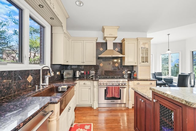 kitchen featuring wall chimney range hood, light wood-type flooring, appliances with stainless steel finishes, cream cabinetry, and a sink