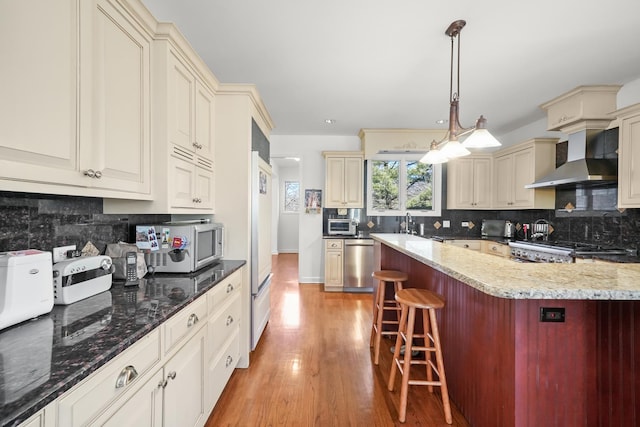 kitchen featuring cream cabinets, dark stone counters, appliances with stainless steel finishes, a breakfast bar area, and wall chimney range hood