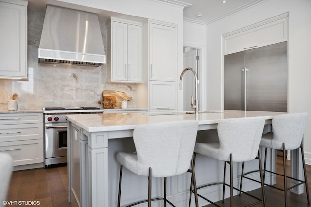 kitchen with high end appliances, dark wood-style floors, a center island with sink, a sink, and wall chimney range hood