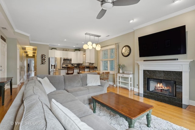 living area with a ceiling fan, baseboards, a high end fireplace, light wood-style floors, and crown molding