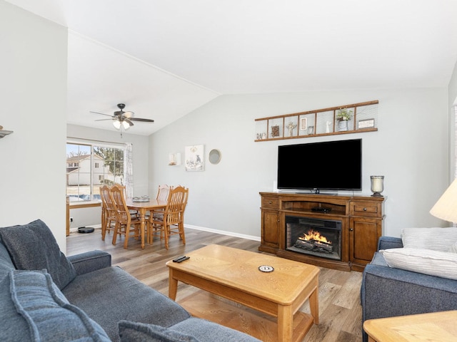 living area featuring baseboards, a warm lit fireplace, ceiling fan, vaulted ceiling, and light wood-style floors