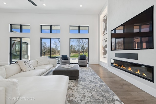 living room featuring plenty of natural light, a towering ceiling, a glass covered fireplace, and dark wood-style floors