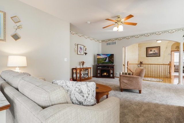 carpeted living room with arched walkways, visible vents, a warm lit fireplace, and a ceiling fan