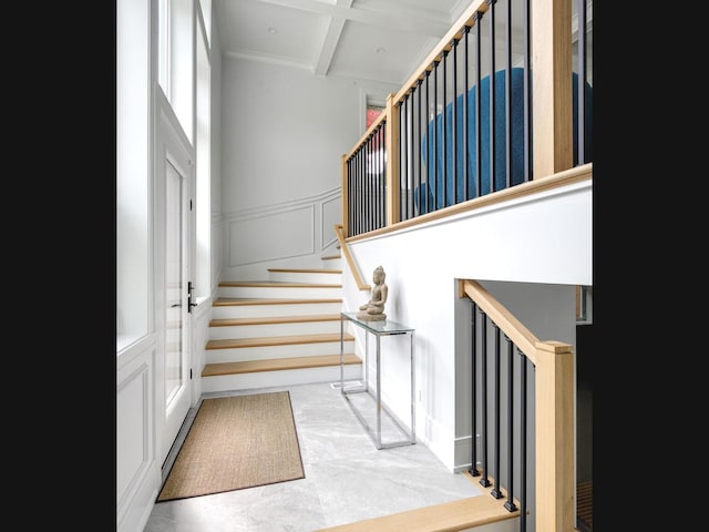 stairs with a wainscoted wall, beamed ceiling, coffered ceiling, a high ceiling, and a decorative wall