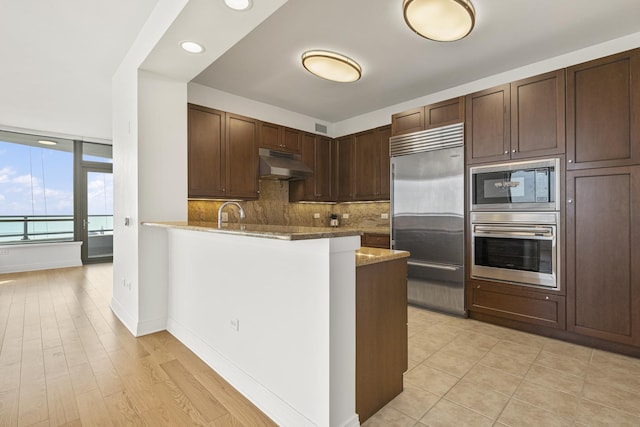 kitchen with a peninsula, dark brown cabinetry, under cabinet range hood, built in appliances, and backsplash