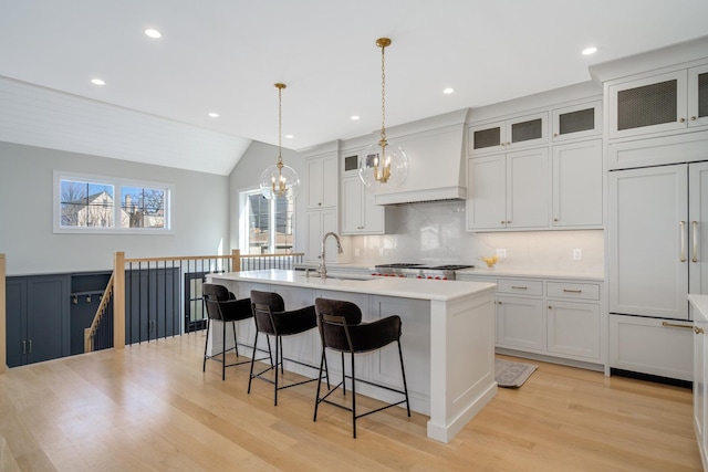 kitchen featuring a center island with sink, custom exhaust hood, a sink, light countertops, and a kitchen bar