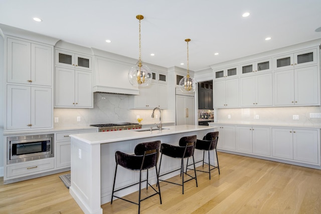 kitchen featuring light wood-type flooring, a center island with sink, a sink, light countertops, and built in appliances