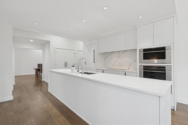kitchen with a sink, double oven, white cabinetry, stovetop, and a kitchen island with sink
