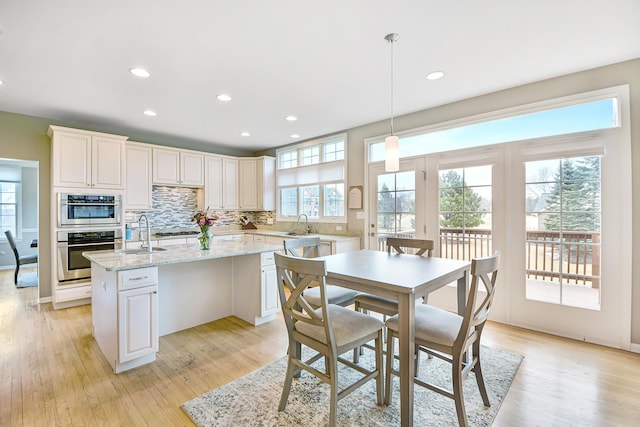 kitchen with light wood finished floors, an island with sink, a sink, double oven, and backsplash