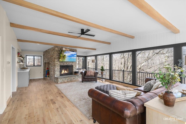 living room featuring beam ceiling, light wood-style floors, a stone fireplace, and a ceiling fan