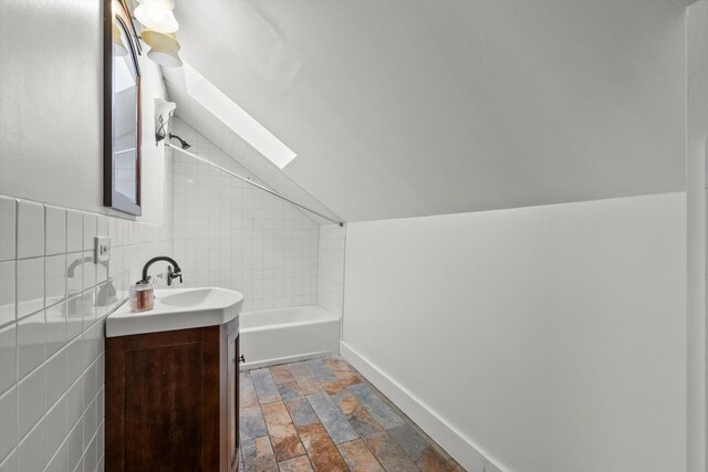 full bathroom featuring tile walls, baseboards, lofted ceiling with skylight, stone finish floor, and vanity