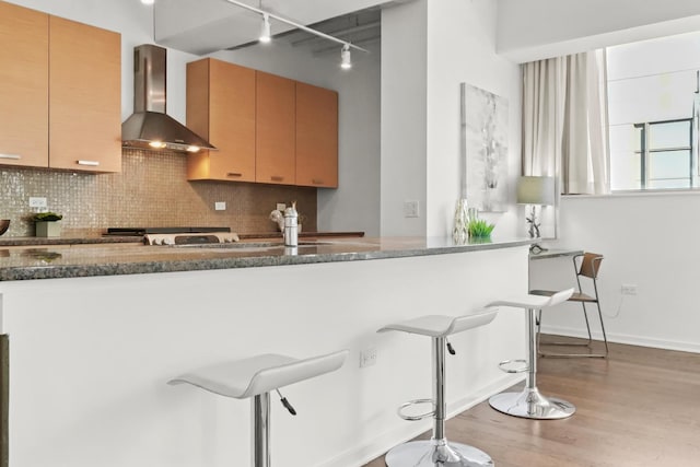 kitchen with a kitchen breakfast bar, tasteful backsplash, wood finished floors, dark stone counters, and wall chimney exhaust hood