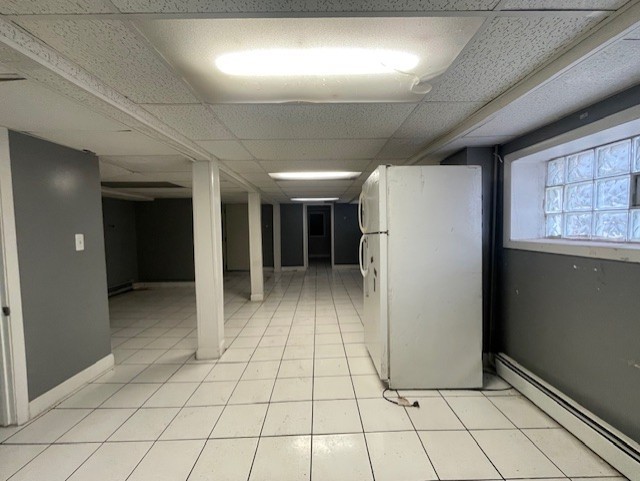 basement with light tile patterned floors, baseboards, a drop ceiling, freestanding refrigerator, and a baseboard heating unit