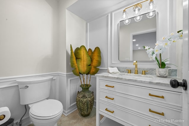 half bathroom with tile patterned flooring, a wainscoted wall, toilet, a decorative wall, and vanity