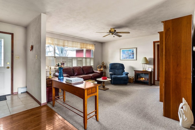 living area with a baseboard heating unit, a glass covered fireplace, baseboards, and ceiling fan