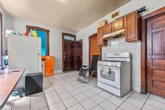 kitchen with brown cabinets, under cabinet range hood, white gas range oven, light tile patterned floors, and decorative backsplash