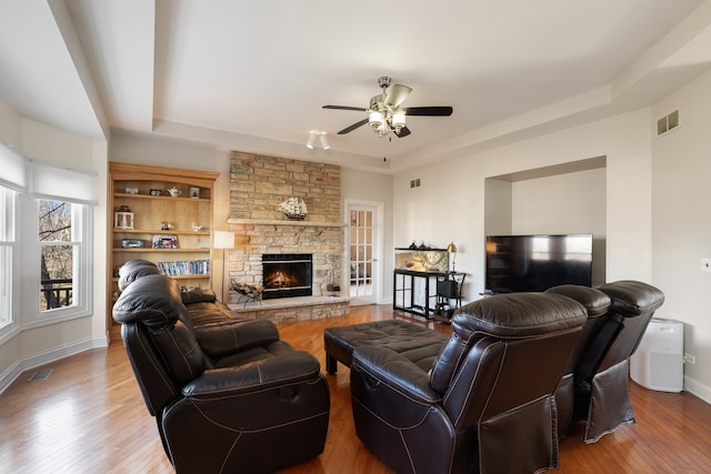 living room featuring wood finished floors, visible vents, baseboards, a stone fireplace, and a raised ceiling