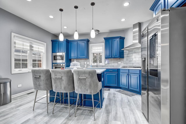 kitchen with blue cabinetry, stainless steel appliances, wall chimney exhaust hood, light countertops, and decorative backsplash