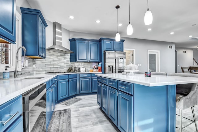 kitchen with a kitchen bar, blue cabinetry, a sink, appliances with stainless steel finishes, and wall chimney range hood