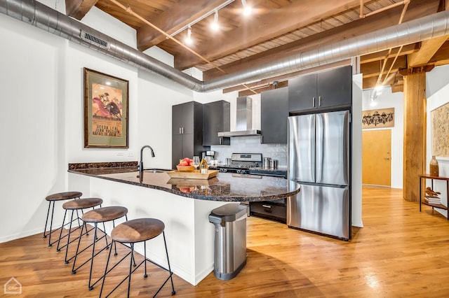 kitchen featuring wall chimney range hood, stainless steel appliances, decorative backsplash, a kitchen breakfast bar, and dark cabinets