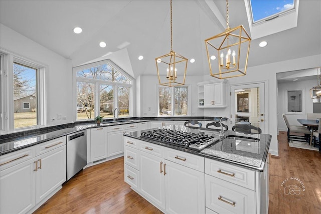 kitchen with dark stone countertops, a kitchen island with sink, light wood-style floors, appliances with stainless steel finishes, and lofted ceiling with skylight