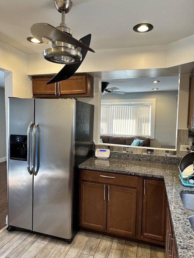 kitchen with wood finish floors, stainless steel fridge with ice dispenser, dark stone counters, a peninsula, and a ceiling fan