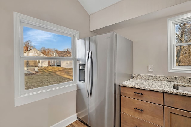 kitchen with light stone counters, a sink, brown cabinetry, stainless steel fridge with ice dispenser, and baseboards