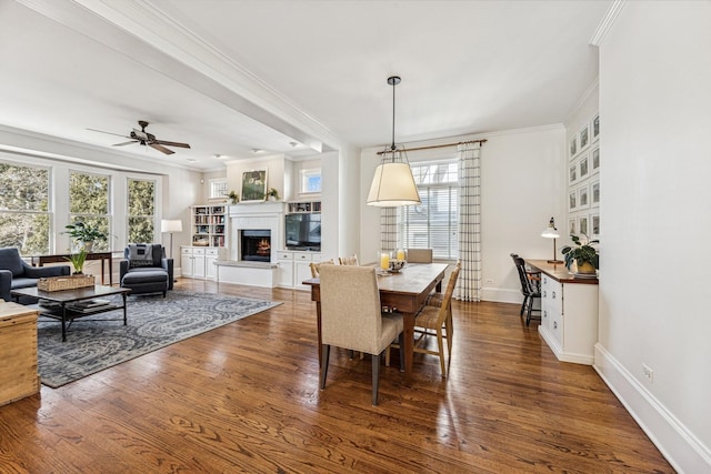 dining space featuring ornamental molding, a ceiling fan, a warm lit fireplace, wood finished floors, and baseboards