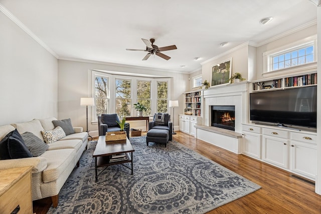 living room with wood finished floors, visible vents, a fireplace, ornamental molding, and ceiling fan