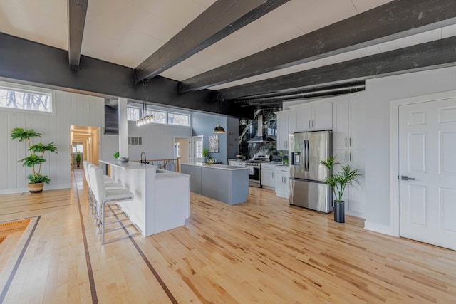 kitchen featuring beamed ceiling, stainless steel appliances, a peninsula, wall chimney exhaust hood, and light wood finished floors