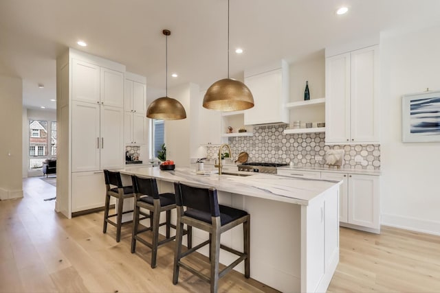 kitchen featuring tasteful backsplash, open shelves, a center island with sink, light wood-type flooring, and a sink