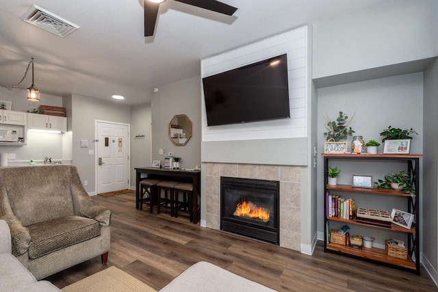 living area featuring a ceiling fan, wood finished floors, visible vents, baseboards, and a tiled fireplace