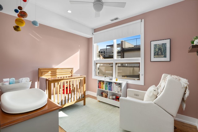 bedroom featuring visible vents, baseboards, a nursery area, wood finished floors, and a ceiling fan