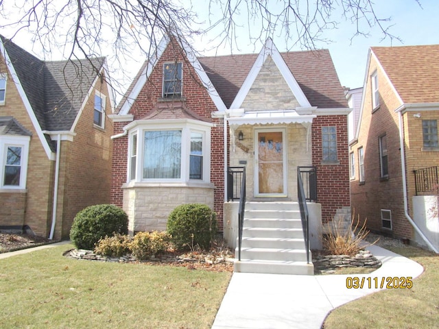 tudor house featuring a front yard, brick siding, stone siding, and roof with shingles