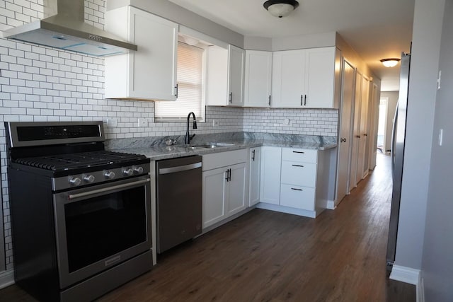 kitchen featuring wall chimney range hood, light stone countertops, white cabinets, stainless steel appliances, and a sink