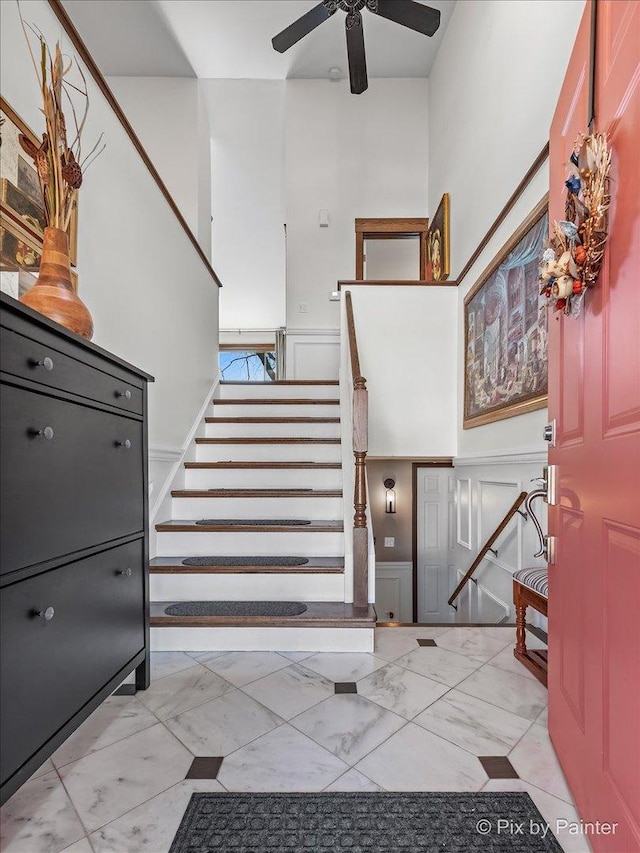 stairs with a ceiling fan, marble finish floor, wainscoting, and a decorative wall