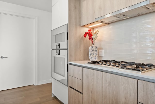 kitchen with light brown cabinets, wood finished floors, stainless steel gas stovetop, wall chimney exhaust hood, and light countertops