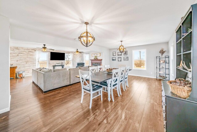 dining room featuring baseboards, wood finished floors, a fireplace, and ceiling fan with notable chandelier