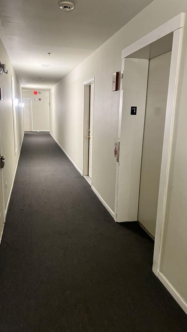 hall with elevator, dark carpet, and baseboards