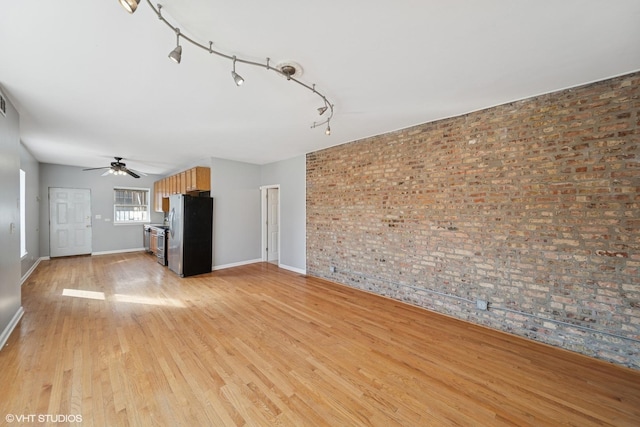 unfurnished living room with light wood finished floors, brick wall, ceiling fan, baseboards, and rail lighting