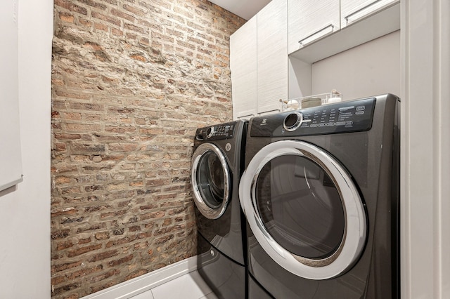 clothes washing area with brick wall, baseboards, washer and dryer, tile patterned floors, and cabinet space