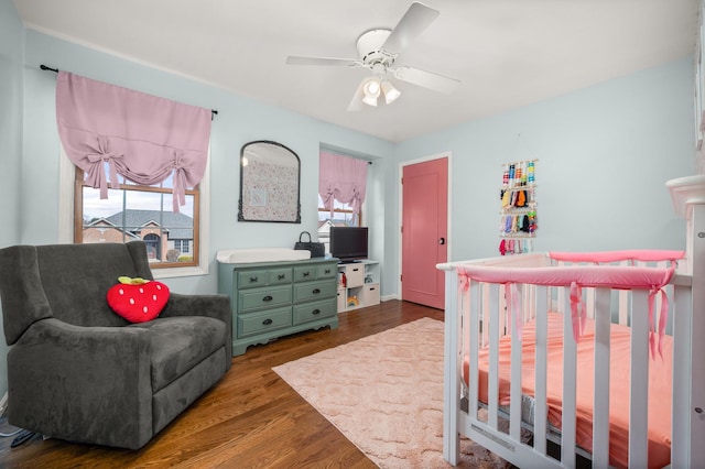 bedroom with dark wood-type flooring, a nursery area, and a ceiling fan