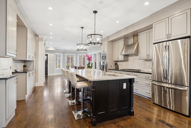 kitchen featuring a center island with sink, dark wood finished floors, a sink, appliances with stainless steel finishes, and wall chimney exhaust hood