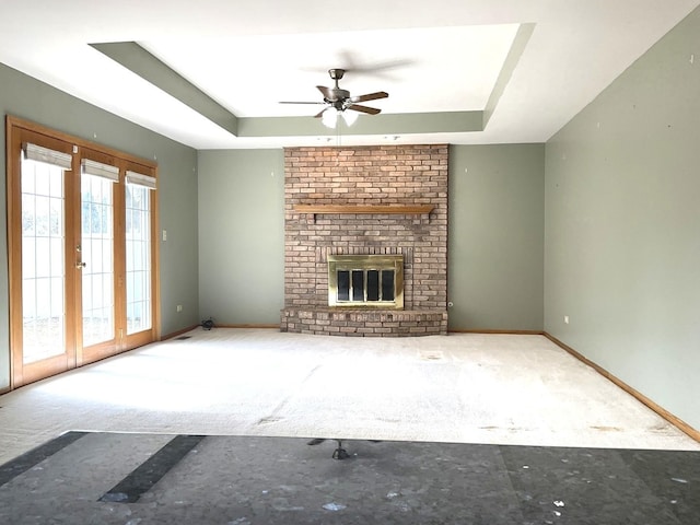 unfurnished living room featuring a brick fireplace, baseboards, a tray ceiling, carpet floors, and a ceiling fan