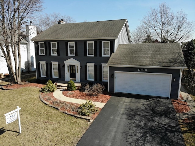 colonial inspired home featuring an attached garage, a shingled roof, a front yard, a chimney, and driveway