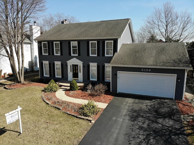 colonial-style house featuring aphalt driveway, a chimney, a garage, and roof with shingles