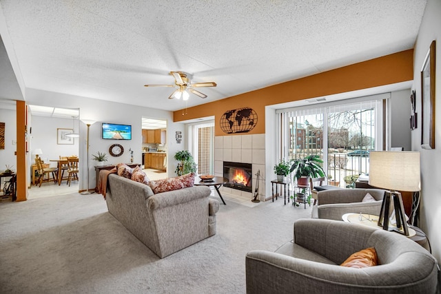 living room with a tiled fireplace, a textured ceiling, a ceiling fan, and light carpet