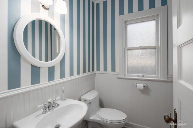 bathroom with a wainscoted wall, toilet, wallpapered walls, and a sink