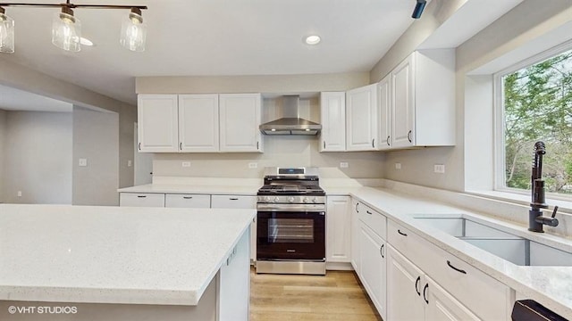 kitchen with wall chimney range hood, stainless steel range with gas cooktop, a sink, white cabinets, and light wood finished floors