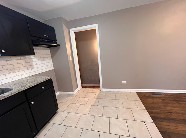 kitchen featuring baseboards, under cabinet range hood, light stone counters, decorative backsplash, and dark cabinets